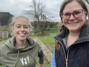 Two women (Mariken on the left, Antonia on the right) smiling broadly. In the background, the plot of a bare allotment is visible.